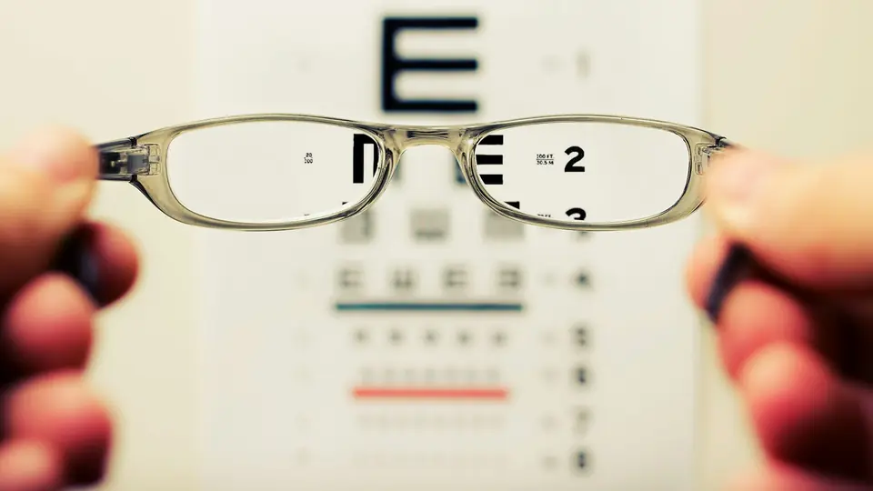 Hands holding a pair of glasses in front of an eye chart